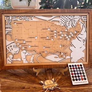 May include: A framed wooden map of the United States featuring laser-cut baseball stadium locations with circular cutouts. The design includes baseball-themed illustrations, a separate color-coded legend card, and a small wooden baseball bat emblem.