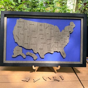 May include: A framed wooden map of the United States with state names engraved. The map is set against a blue background and framed in black. Several detached wooden state pieces are at the bottom. The map is displayed on a wooden surface.