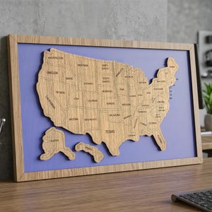 May include: A wooden map of the United States with state outlines and names engraved. The map is set against a light purple background and framed in natural wood. The states of Alaska and Hawaii are separate pieces.