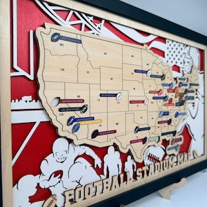 Könnte beinhalten: Ein mehrschichtiges Wandkunstwerk aus Holz mit einer Karte der Vereinigten Staaten, Staatskürzeln und footballförmigen Markierungen für Stadionstandorte. Das Design umfasst einen roten Hintergrund, weiße Silhouetten von Footballspielern und den Schriftzug FOOTBALL STADIUM MAP.