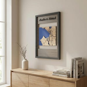 May include: A framed wooden map with the text "Amelia & Patrick" at the top. The map shows a blue water area and a detailed beige land area, set within a grey frame. The artwork is displayed on a light-coloured wall.
