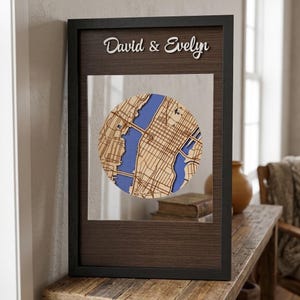May include: A framed wooden map of a city, with a circular design showing blue water and brown land. The black frame has the names "David & Evelyn" in white script. The map is displayed on a wooden surface, with a dark brown border.