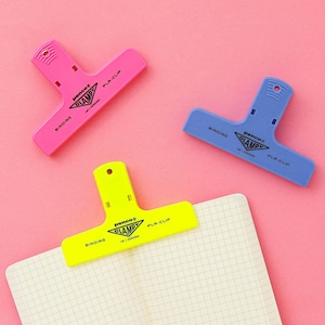 Puede incluir: Tres clips de encuadernaci&oacute;n de pl&aacute;stico de colores rosa, azul y amarillo, cada uno con el texto "BINDING", "CLAMP" y "PLA-CLIP". Un clip est&aacute; sujeto a un cuaderno con un patr&oacute;n de cuadr&iacute;cula, todo sobre un fondo rosa.