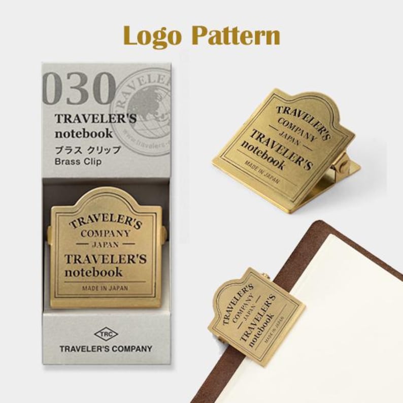 May include: A brass clip for a Traveler's notebook, with the text "TRAVELER'S COMPANY - JAPAN - TRAVELER'S notebook MADE IN JAPAN". The clip is shown in a box and attached to a notebook. The box has the text "030 TRAVELER'S notebook Brass Clip".