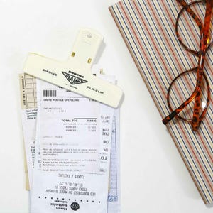 Puede incluir: Un clip Penco Clampy PLA-CLIP color crema sujetando recibos y una postal. El clip tiene las palabras "BINDING" y "CLAMPY" impresas. Tambi&eacute;n se ven unas gafas carey y un cuaderno a rayas.