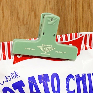 Puede incluir: Un clip de bolsa de pl&aacute;stico verde claro, con la marca "penco CLAMPY PLA-CLIP", est&aacute; sujeto a una bolsa con rayas rojas y blancas. La bolsa tiene texto japon&eacute;s y letras azules. El clip es de pl&aacute;stico resistente.