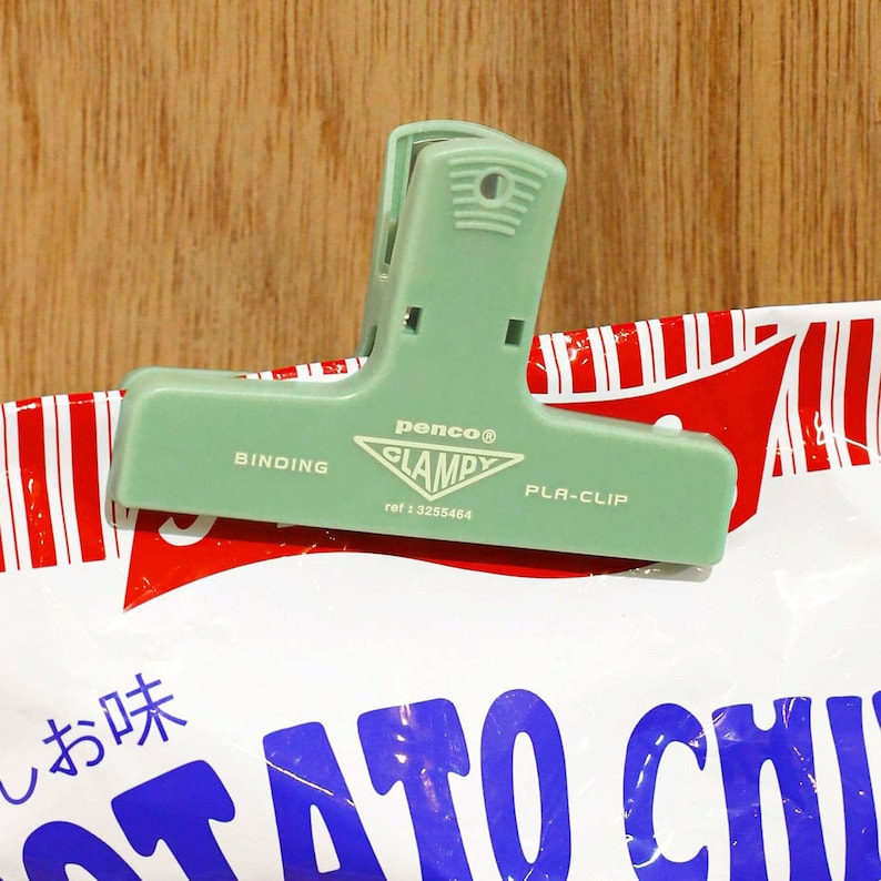 Puede incluir: Un clip de bolsa de pl&aacute;stico verde claro, con la marca "penco CLAMPY PLA-CLIP", est&aacute; sujeto a una bolsa con rayas rojas y blancas. La bolsa tiene texto japon&eacute;s y letras azules. El clip es de pl&aacute;stico resistente.