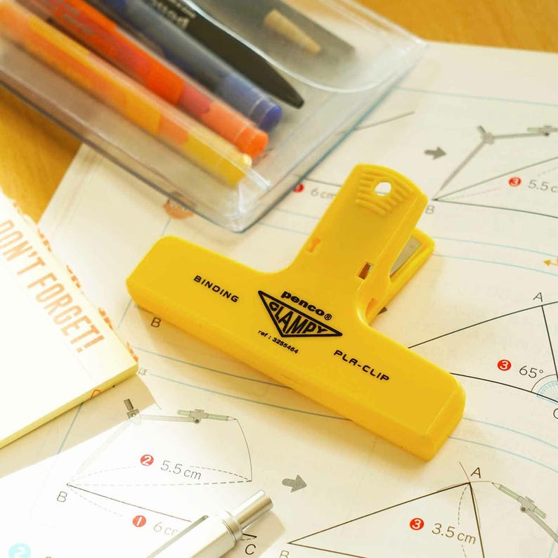 Puede incluir: Un clip amarillo "penco CLAMPY PLA-CLIP" descansa sobre una p&aacute;gina con diagramas geom&eacute;tricos. El clip tiene la palabra "BINDING" impresa. Tambi&eacute;n se ve una funda de pl&aacute;stico transparente con bol&iacute;grafos y un bloc de notas con el texto "DON'T FORGET!"