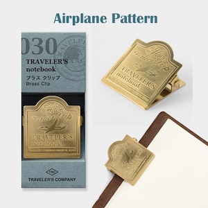 May include: Brass clip for a Traveler's notebook with an airplane design. The clip is gold-colored and features the text "Have a nice trip" and "Traveler's notebook". The product is packaged in a blue box with the text "Airplane Pattern".