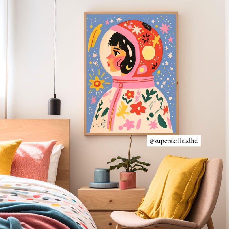 May include: A colorful illustration of a person wearing a pink and red astronaut suit with flowers and stars on it. The background is blue with white stars. The illustration is framed in a wooden frame. The text "@superskillsadhd" is at the bottom of the image.