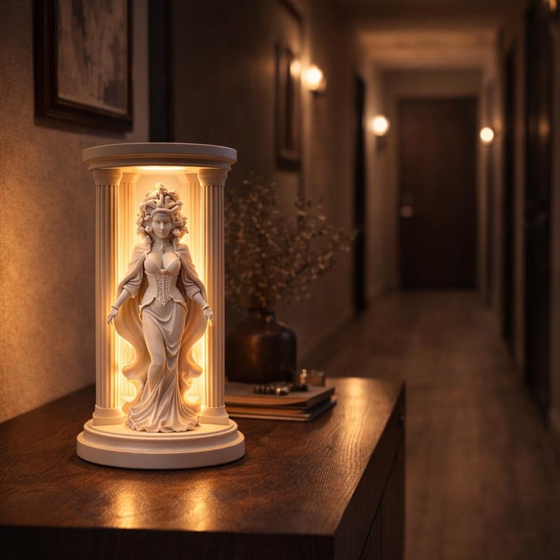 Puede incluir: Una estatua blanca e iluminada de una mujer con un vestido cl&aacute;sico, situada dentro de una estructura similar a una columna. La estatua est&aacute; iluminada desde dentro, creando un brillo c&aacute;lido. La l&aacute;mpara est&aacute; sobre una superficie de madera oscura.