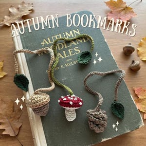 Peut inclure: Trois marque-pages au crochet avec des motifs sur le thème de l'automne : un gland beige, un champignon rouge et blanc, et une pomme de pin marron. Chaque marque-page possède un long cordon fin terminé par une petite feuille vert foncé.