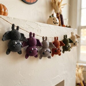 May include: A garland featuring six crocheted bats in various colors including dark gray, purple, light gray, orange, olive green, and cream. Each bat has black bead eyes, small white fangs, and is suspended from a thin brown twine string.