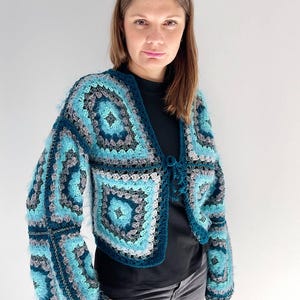 Può includere: Una giacca all'uncinetto turchese, grigia e blu con un motivo quadrato. La giacca ha maniche lunghe e una chiusura con lacci sul davanti. L'uncinetto ha un aspetto strutturato e soffice.