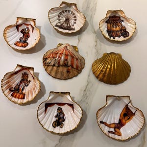 Puede incluir: Colección de conchas de vieira decorativas, cada una con temática de circo. Las conchas tienen un interior blanco con ribete dorado y presentan imágenes relacionadas con el circo, como una noria, un tigre y un oso en bicicleta.