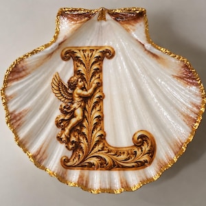 May include: A decorative scallop shell dish featuring a large, ornate letter L in brown and gold tones. The letter is integrated with a winged cherub figure and intricate scrollwork. The shell has a white surface with brown accents and a gold-painted rim.