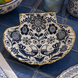 Può includere: Un piatto decorativo a forma di conchiglia con base bianca e motivo floreale e di uccelli blu. Il bordo è rifinito con oro. Il piatto è su una superficie piastrellata blu, con altre stoviglie blu e bianche visibili.