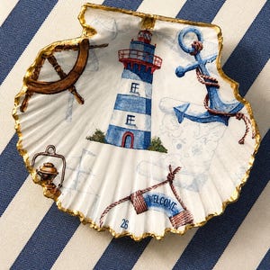 Può includere: Un piatto decorativo a forma di conchiglia con bordo dorato. L'interno presenta un'illustrazione nautica con un faro blu e bianco, un timone, un'ancora blu con corda, una lanterna e uno stendardo con la scritta WELCOME.