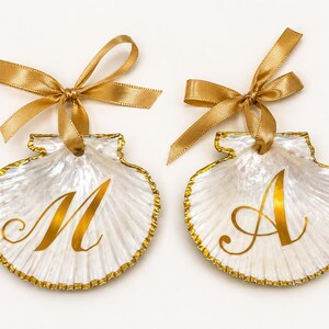 May include: Two white scallop shell ornaments featuring gold-painted edges and a gold ribbon bow at the top. Each shell displays a large, elegant gold letter in the center, with one shell featuring the letter M and the other featuring the letter A.