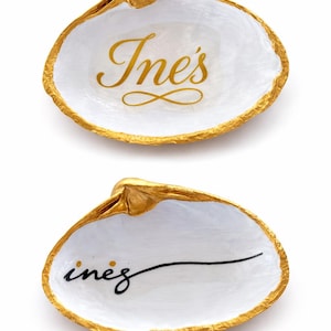 May include: Two white seashell dishes with gold-painted edges. The top shell features the name Ine's in gold script with an infinity symbol below. The bottom shell features the name inez in black cursive script with a long horizontal underline.