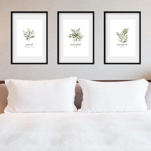 May include: Three framed prints with black frames and white mounts. Each print features a watercolour illustration of a sprig of greenery with the name of the plant written below. The prints are hung on a white wall above a white bed with white bedding.