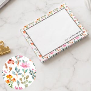 May include: A rectangular notepad featuring a watercolor floral border with pink, orange, and green botanical elements. The center is white with a thin gray border and the printed text Mrs. Caroline Smith. A gold metal binder clip is positioned nearby.