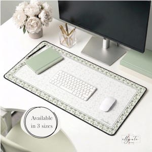 Puede incluir: Una alfombrilla de escritorio rectangular con superficie blanca y un delicado borde con estampado floral verde. La alfombrilla incluye un teclado de ordenador blanco, un ratón blanco y un cuaderno verde salvia. El nombre Caroline está impreso en cursiva.
