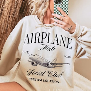 May include: Cream-colored sweatshirt with the text "AIRPLANE Mode Social Club" and an airplane graphic. The sweatshirt also has the text "EST. 2026" and "CUSTOM LOCATION". The sweatshirt is a casual style.