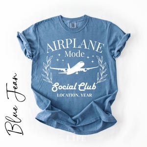 May include: A blue jean-colored t-shirt with the words "AIRPLANE Mode Social Club" in white, along with an airplane graphic. The shirt has rolled-up sleeves and is displayed on a white background. The words "Blue Jean" are written in cursive on the left side.