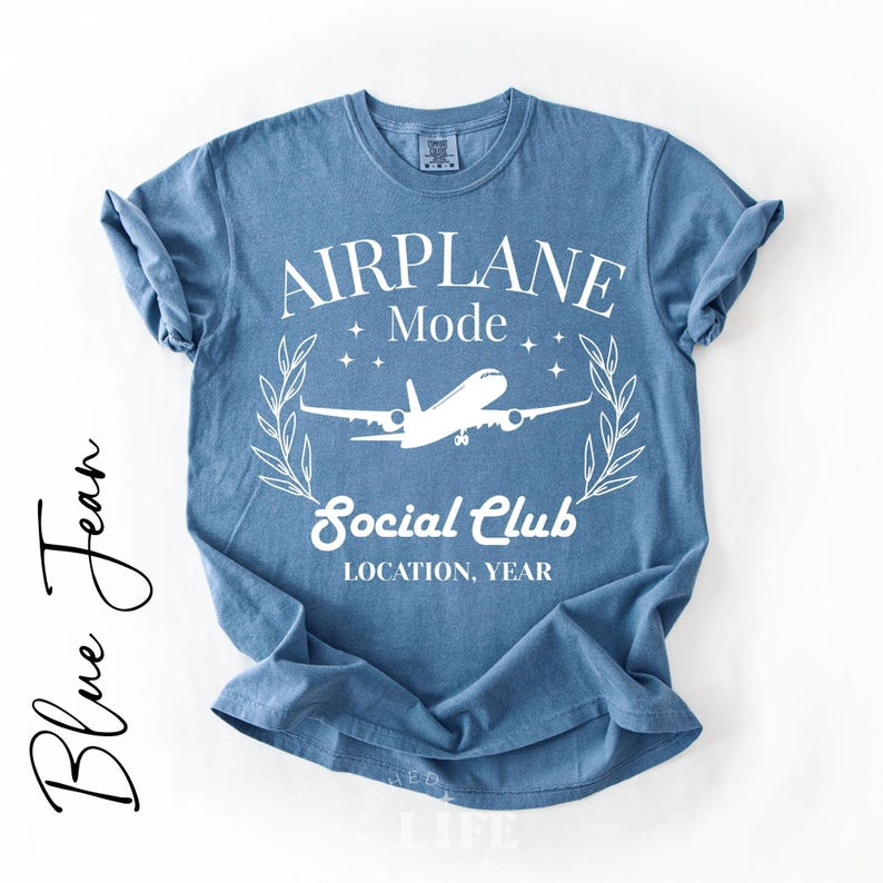May include: A blue jean-colored t-shirt with the words "AIRPLANE Mode Social Club" in white, along with an airplane graphic. The shirt has rolled-up sleeves and is displayed on a white background. The words "Blue Jean" are written in cursive on the left side.