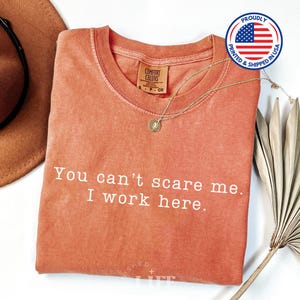 May include: A coral-colored t-shirt with the text "You can't scare me. I work here." printed in white. The shirt is made of a soft, comfortable material and has a distressed look.