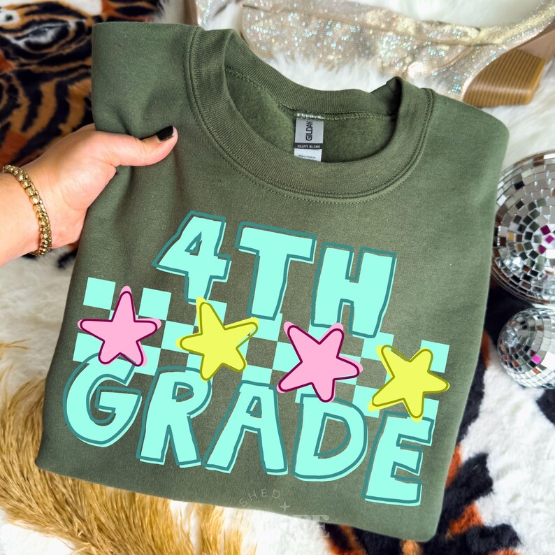May include: Olive green sweatshirt with the words "4TH GRADE" in light blue, outlined in white, with pink and yellow stars. The text is over a checkered pattern. A gold bracelet is visible.