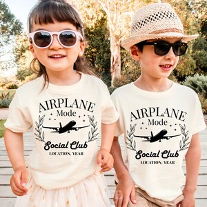 May include: Two children wearing cream-colored t-shirts with the text "AIRPLANE Mode Social Club" and an airplane graphic. The shirts have a black design. One child wears pink sunglasses, the other a straw hat and black sunglasses.