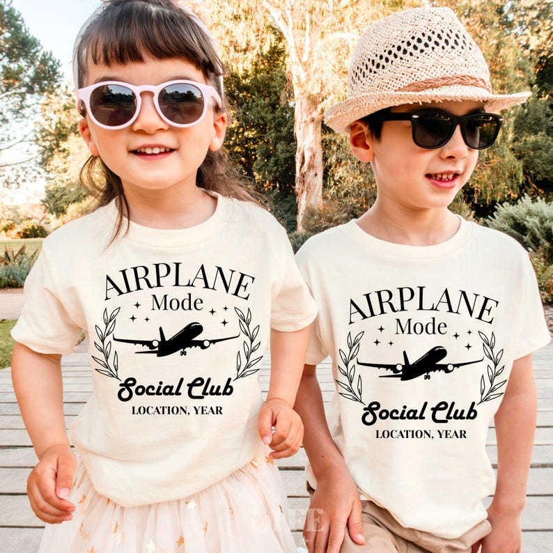 May include: Two children wearing cream-colored t-shirts with the text "AIRPLANE Mode Social Club" and an airplane graphic. The shirts have a black design. One child wears pink sunglasses, the other a straw hat and black sunglasses.