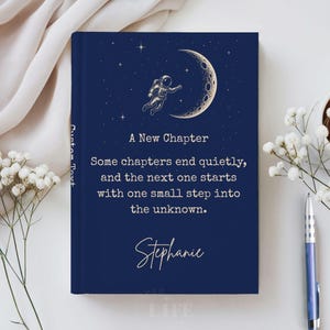 May include: A dark blue hardcover journal featuring a gold illustration of an astronaut floating near a crescent moon and stars. The text reads: A New Chapter, Some chapters end quietly, and the next one starts with one small step into the unknown. Stephanie.