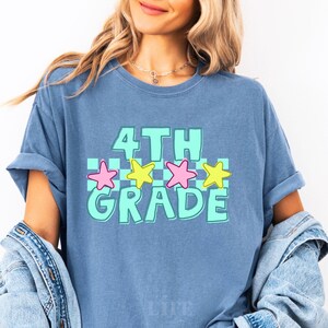 May include: A light blue t-shirt with the words "4TH GRADE" in a turquoise and white checkered design. The shirt features pink and yellow star graphics. The shirt is made of a soft material.