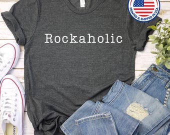 Puede incluir: Una camiseta gris oscuro con el texto "Rockaholic" impreso en blanco en la parte delantera. La camiseta está colocada sobre una superficie de madera con un par de pantalones vaqueros azules y zapatillas blancas.