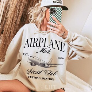 Puede incluir: Sudadera color crema con el texto "AIRPLANE Mode Social Club" y un gr&aacute;fico de avi&oacute;n. La sudadera tambi&eacute;n tiene el texto "EST. 2026" y "CUSTOM LOCATION". El estilo es informal.