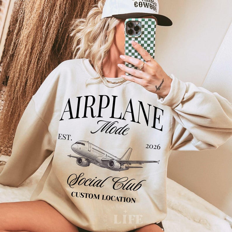 Puede incluir: Sudadera color crema con el texto "AIRPLANE Mode Social Club" y un gr&aacute;fico de avi&oacute;n. La sudadera tambi&eacute;n tiene el texto "EST. 2026" y "CUSTOM LOCATION". El estilo es informal.