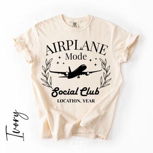 May include: Ivory-colored t-shirt with the words "AIRPLANE Mode Social Club" and a silhouette of an airplane. The design includes stars and a laurel wreath. The text "LOCATION, YEAR" is also included.