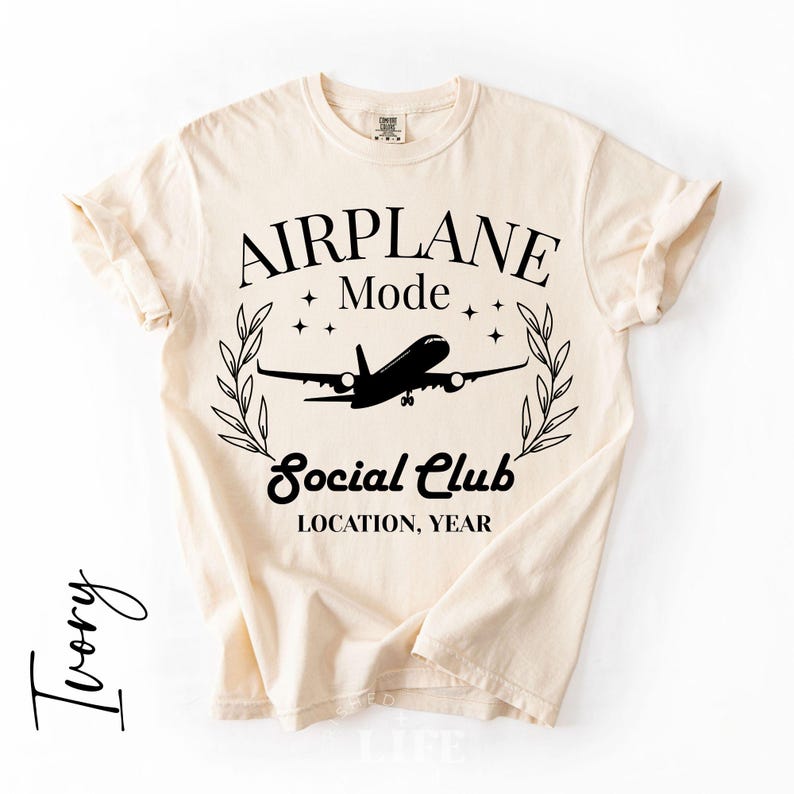 May include: Ivory-colored t-shirt with the words "AIRPLANE Mode Social Club" and a silhouette of an airplane. The design includes stars and a laurel wreath. The text "LOCATION, YEAR" is also included.