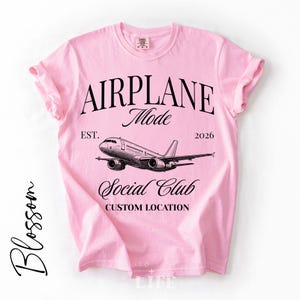 May include: A light pink t-shirt with rolled-up sleeves. The shirt features the text "AIRPLANE Mode Social Club" with an airplane graphic. The text "EST. 2026" and "CUSTOM LOCATION" are also printed on the shirt.