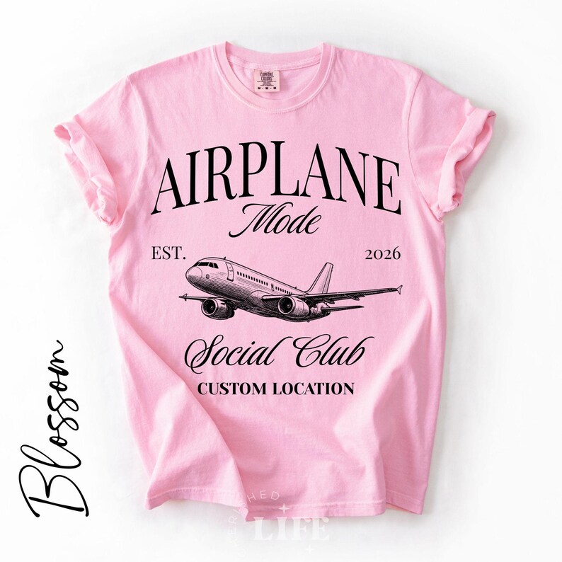 Puede incluir: Camiseta rosa claro con las palabras "AIRPLANE Mode" y un gr&aacute;fico de un avi&oacute;n. El texto "EST. 2026" y "Social Club" tambi&eacute;n est&aacute;n impresos en la camiseta. Las palabras "CUSTOM LOCATION" est&aacute;n impresas debajo de las palabras "Social Club".