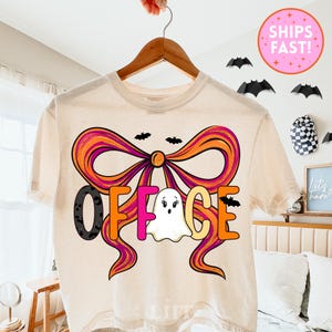 May include: Cream-colored t-shirt with a large bow design in orange and purple, featuring the word "OFFICE" in black, pink, orange, and yellow. A white ghost graphic is in the center. Small black bats are scattered around the design.