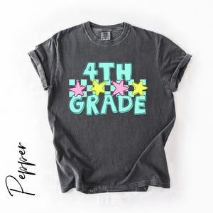 May include: Dark gray t-shirt with the words "4TH GRADE" in a turquoise font, outlined in white. Pink and yellow stars are above the word "GRADE". The shirt has rolled-up sleeves and a crew neck.