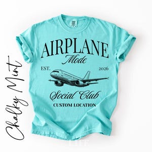 Puede incluir: Una camiseta azul claro con las palabras "AIRPLANE Mode Social Club" y un gr&aacute;fico de avi&oacute;n. La camiseta tambi&eacute;n tiene el texto "EST. 2026" y "CUSTOM LOCATION". La camiseta es de color azul claro liso.