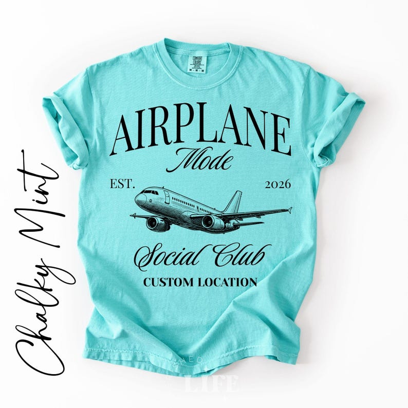 Puede incluir: Una camiseta azul claro con las palabras "AIRPLANE Mode Social Club" y un gr&aacute;fico de avi&oacute;n. La camiseta tambi&eacute;n tiene el texto "EST. 2026" y "CUSTOM LOCATION". La camiseta es de color azul claro liso.
