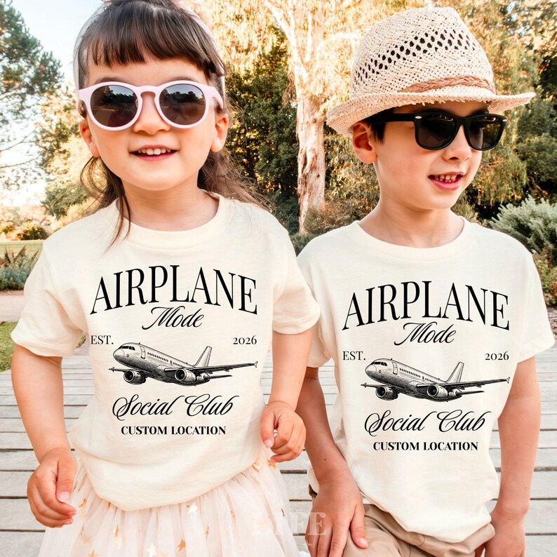 Puede incluir: Dos ni&ntilde;os con camisetas color crema con el texto "AIRPLANE Mode" sobre un gr&aacute;fico de avi&oacute;n. Las camisetas tambi&eacute;n dicen "Social Club" y "Custom Location" con el a&ntilde;o "2026". Uno lleva gafas de sol rosas, el otro un sombrero de paja.