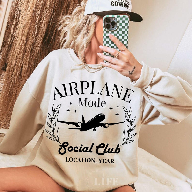 May include: Cream-colored sweatshirt with the text "AIRPLANE Mode Social Club" in black, featuring an airplane graphic. The design includes stars and laurel branches. The sweatshirt has long sleeves.