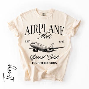 Puede incluir: Camiseta de color marfil con las palabras "AIRPLANE Mode Social Club" y una ilustraci&oacute;n detallada de un avi&oacute;n. La camiseta tambi&eacute;n incluye el texto "EST. 2026" y "CUSTOM LOCATION".